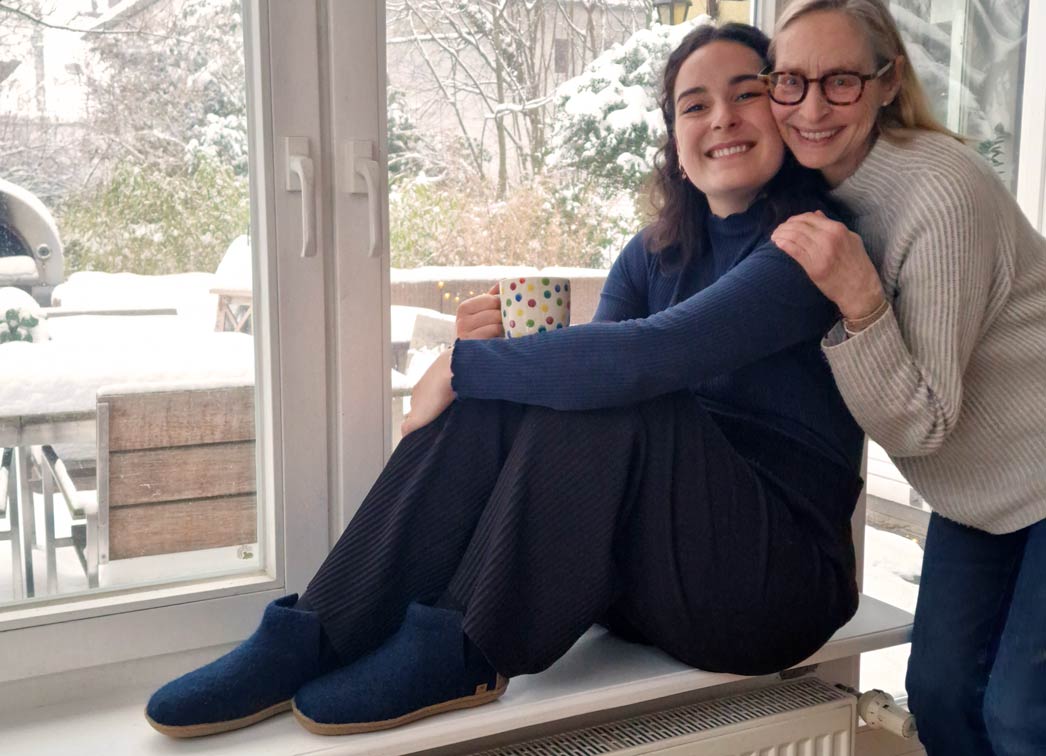 Zwei Frauen schauen lächelnd in die Kamera. Eine trägt die warmen Filzhausschuhe WoolFit Yeti in einem traumhaften Blauton
